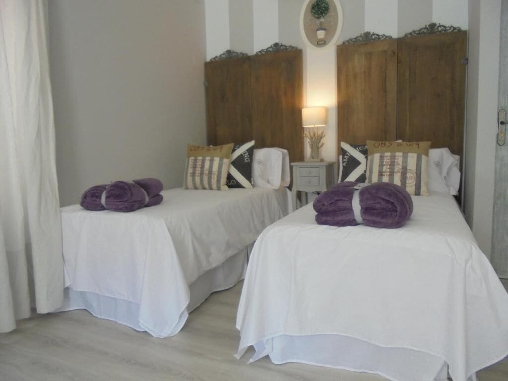 A hotel room with two beds, each with a white bedspread and purple towels. There are wooden headboards and a small bedside table with a lamp.