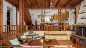 A spacious living area with wooden furniture, a stone fireplace, and a kitchen in the background. - Yanni’s Place (93MF+669 Petaloudes, Greece)