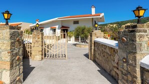 A traditional stone-walled building with a red-tiled roof, a gated entrance, and two street lamps. - Yanni’s Place (93MF+669 Petaloudes, Greece)