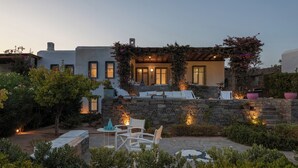A house with a stone wall, a patio with white furniture, and a garden with various plants. - Isterni | 3BR Sea View House | Coral Pearl (Paros)