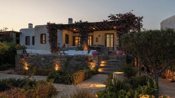 A Mediterranean-style villa with a stone facade, outdoor seating area, and a garden with cacti and shrubs. - Isterni | 3BR Sea View House | Coral Pearl (Paros)