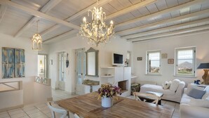 A spacious living area with a wooden dining table, white furniture, a chandelier, and a view of the outdoors through large windows. - Isterni | 3BR Sea View House | Coral Pearl (Paros)