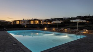 A swimming pool with lounge chairs and umbrellas, surrounded by stone flooring and buildings in the background. - Isterni | 3BR Sea View House | Coral Pearl (Paros)