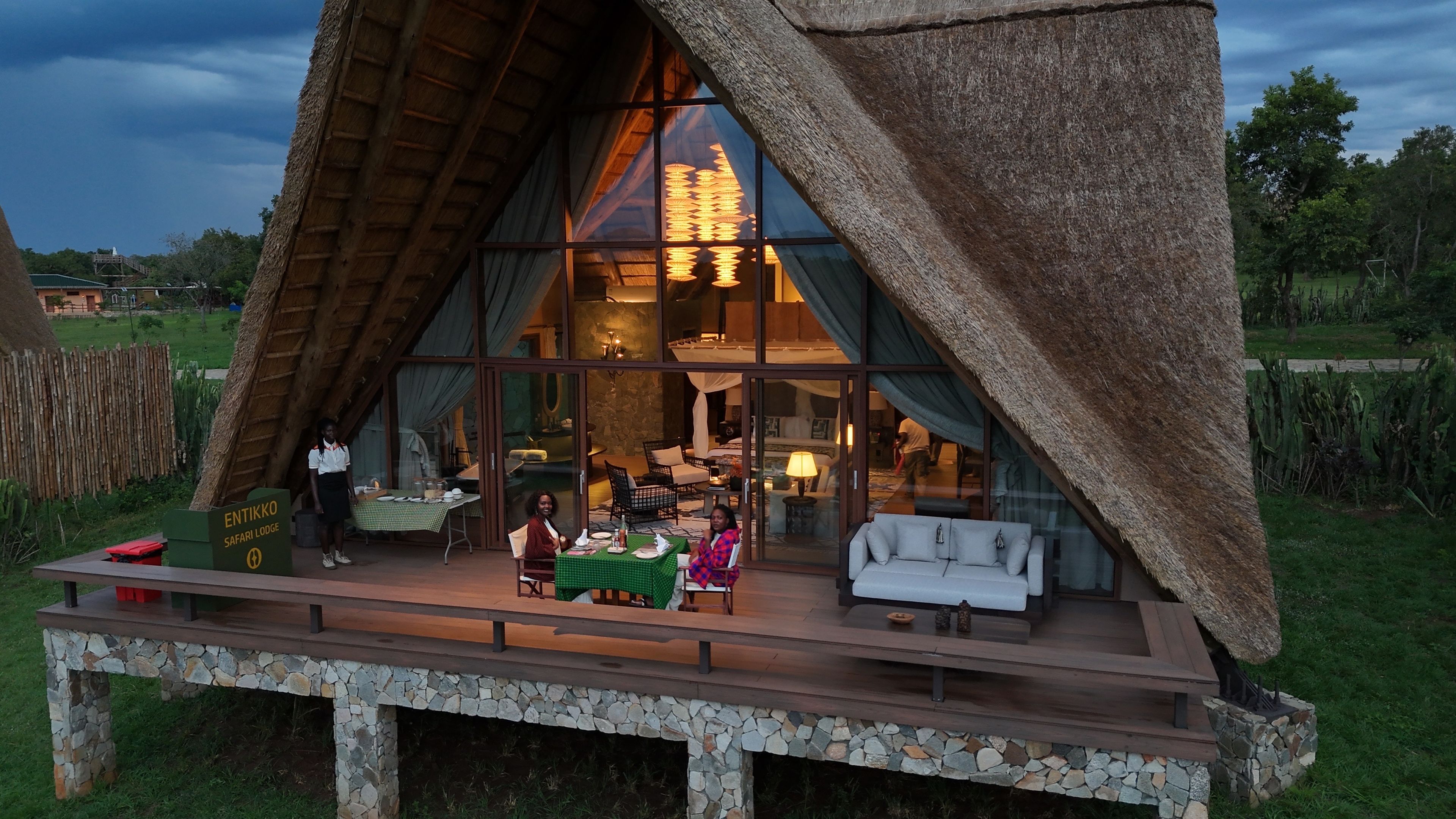 A safari lodge with a thatched roof, large windows, and a stone deck.
