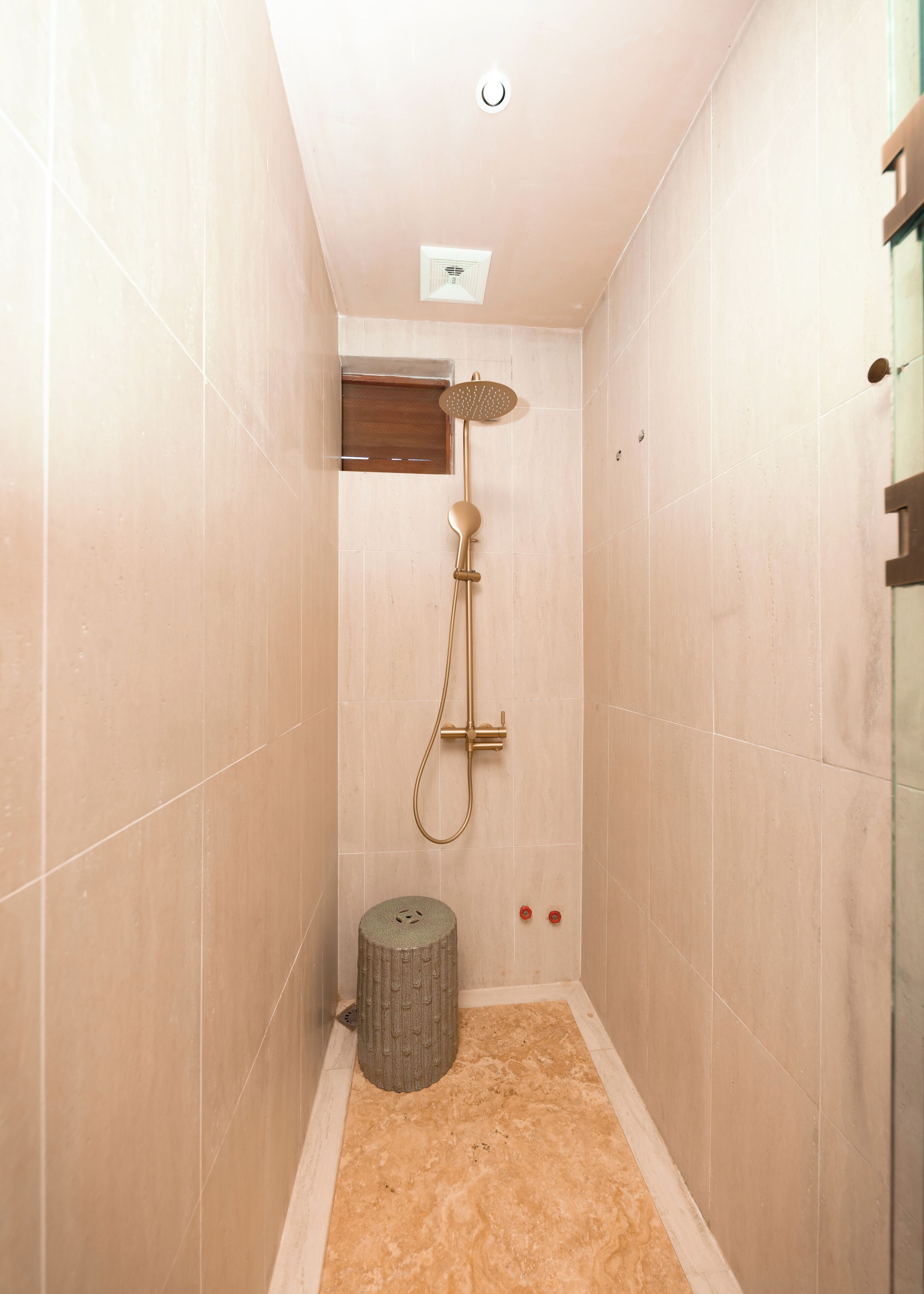 Une petite salle de douche étroite, avec des carreaux de couleur beige, une pomme de douche et un petit tabouret.