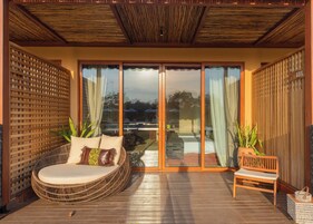 Una terraza de madera con un sofá de mimbre y un banco de madera, amplias puertas correderas de vidrio que dan a una zona de piscina y un techo de madera.