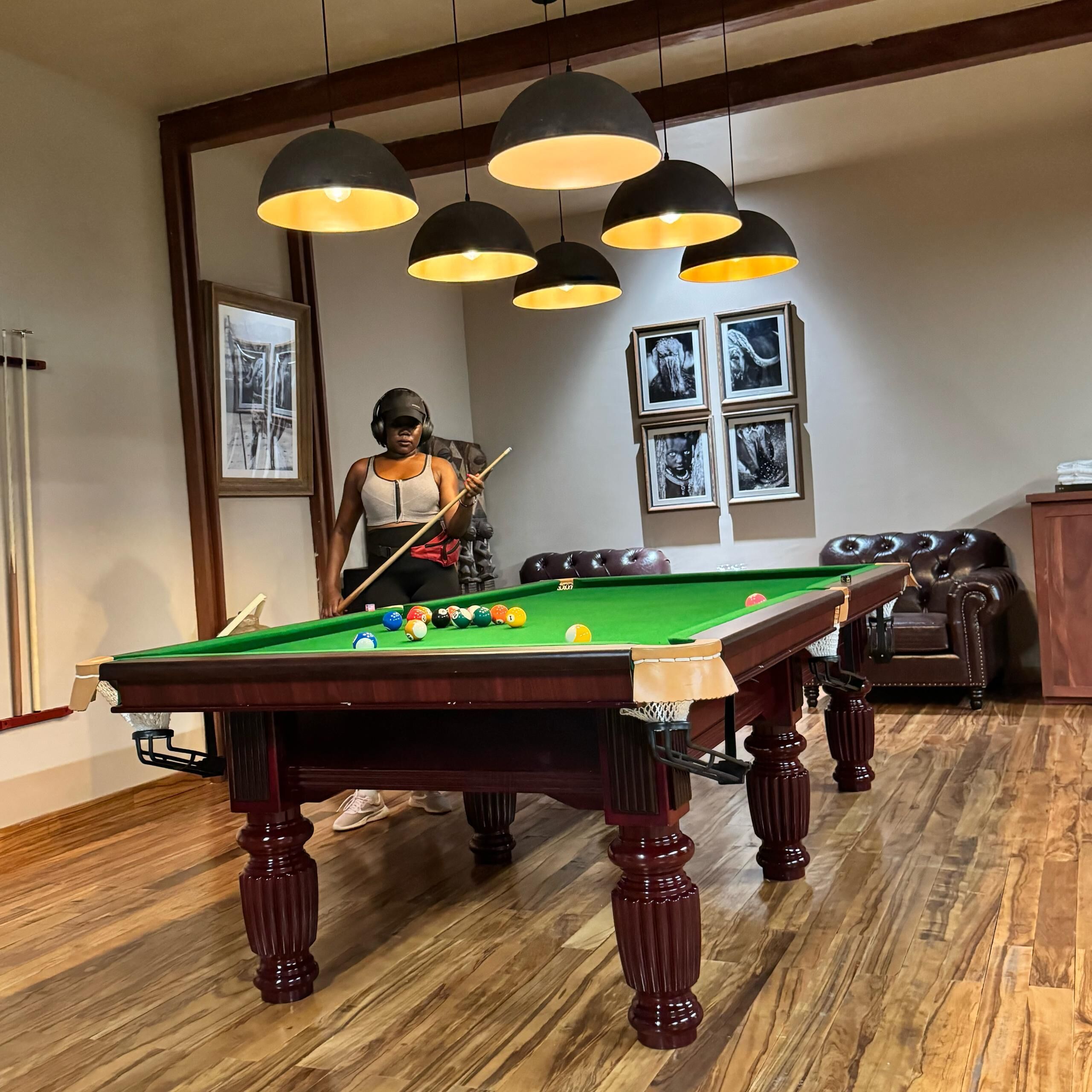 Een biljart tafel in een kamer met houten vloeren, een leren bank en een plafond met hangende lampen.