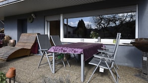 Un patio al aire libre con una mesa y sillas, un banco de madera y una ventana con vista a una casa y árboles.
