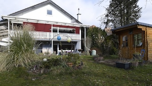 Una casa de dos pisos con balcón, una pequeña construcción de madera y un jardín con plantas variadas y césped.
