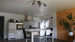Una cocina moderna con armarios blancos, una mesa de comedor de madera y sillas. Hay un microondas, un horno y una planta en el alféizar de la ventana.