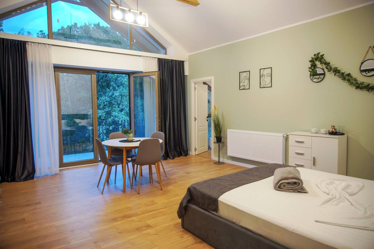 A modern hotel room with a large bed, a dining area, and a view through the glass doors.