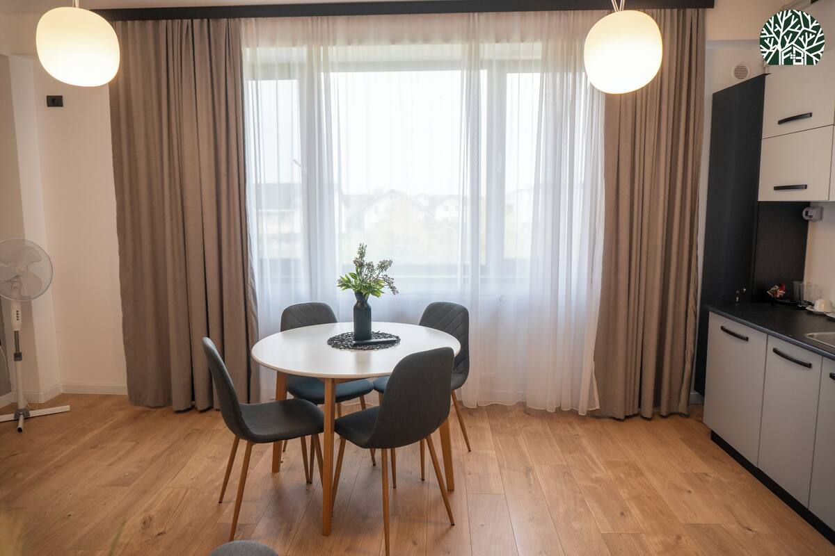 A dining area with a round table, six chairs, a vase with flowers, and a view through large windows with curtains.