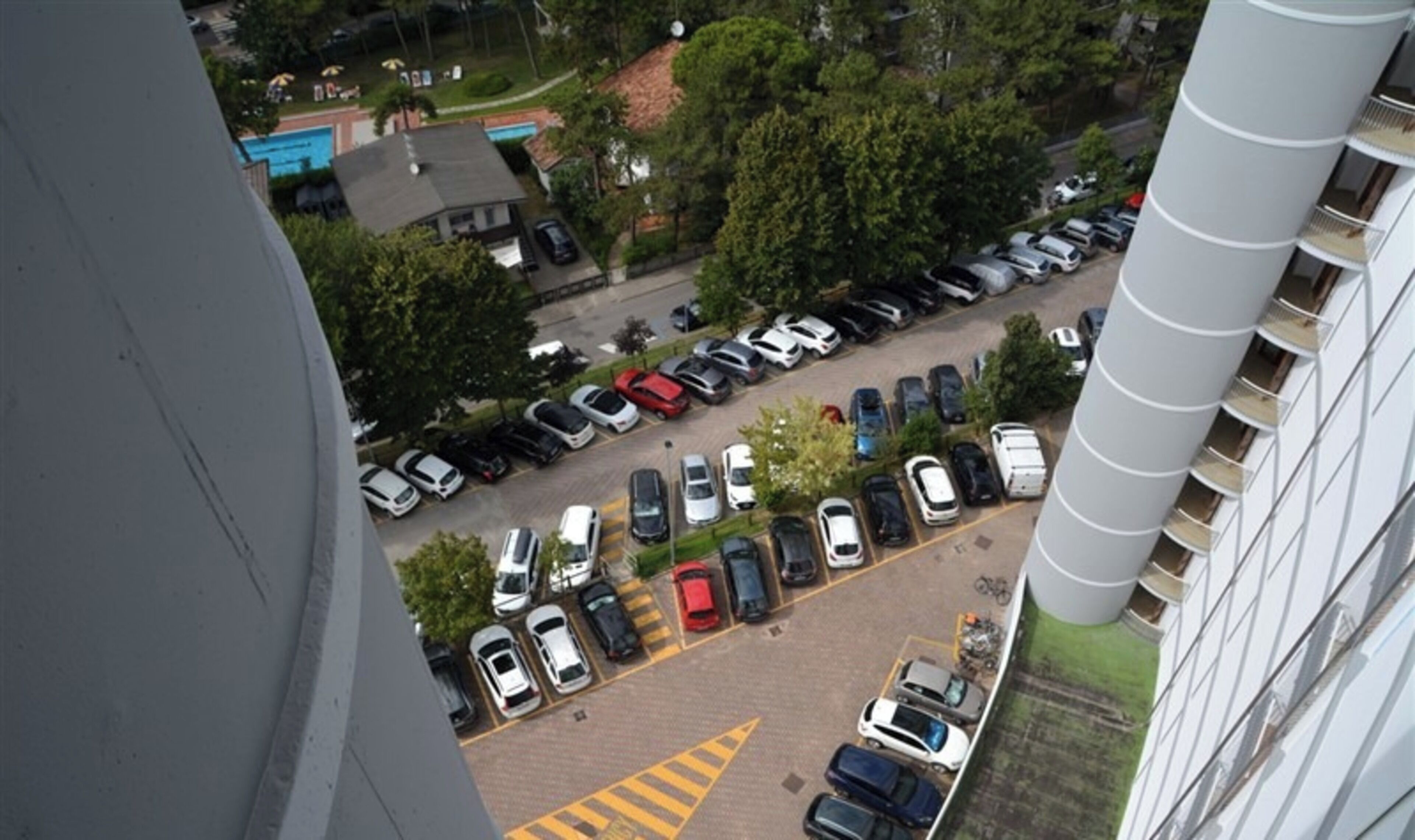 An aerial view of a parking lot with multiple rows of parked cars, surrounded by trees and buildings.