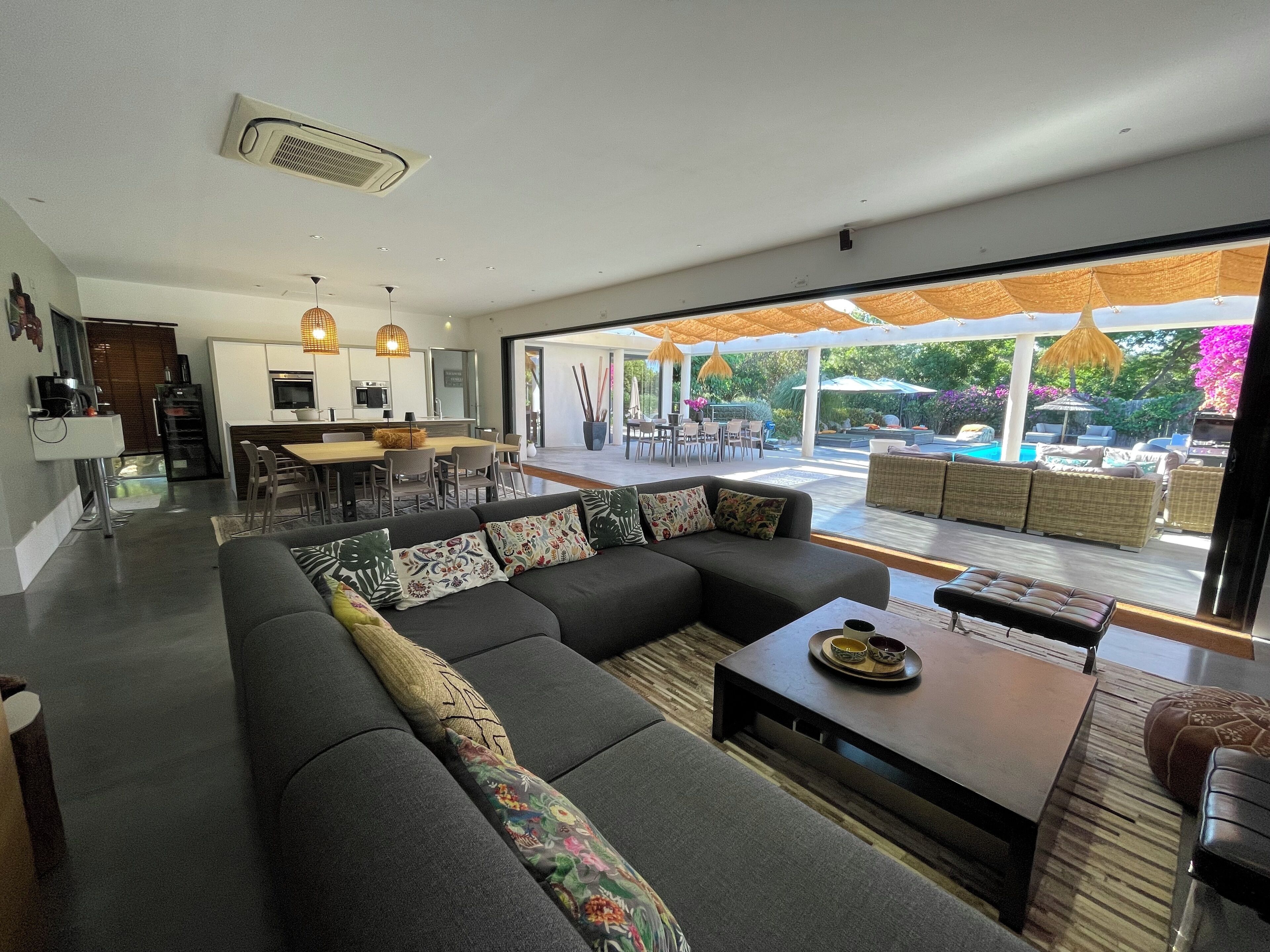 A modern living room with a large sectional sofa, a dining area, and a view of a pool and garden.