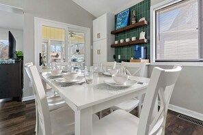 A dining area with a white table set for four, surrounded by white chairs. A green tiled backsplash and a window with blinds are visible.