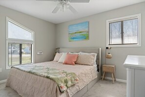A bedroom with a bed, a ceiling fan, a painting, a window with blinds, and a bedside table.