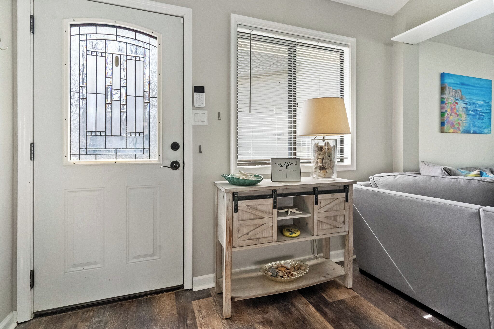 A room with a white door, a window with blinds, a lamp on a wooden console table, and a gray sofa.