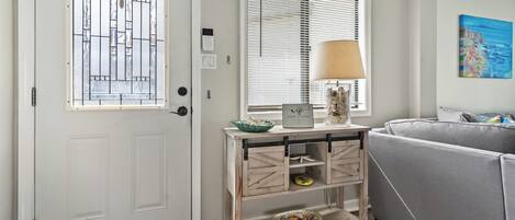 A room with a white door, a window with blinds, a lamp on a wooden console table, and a gray sofa.
