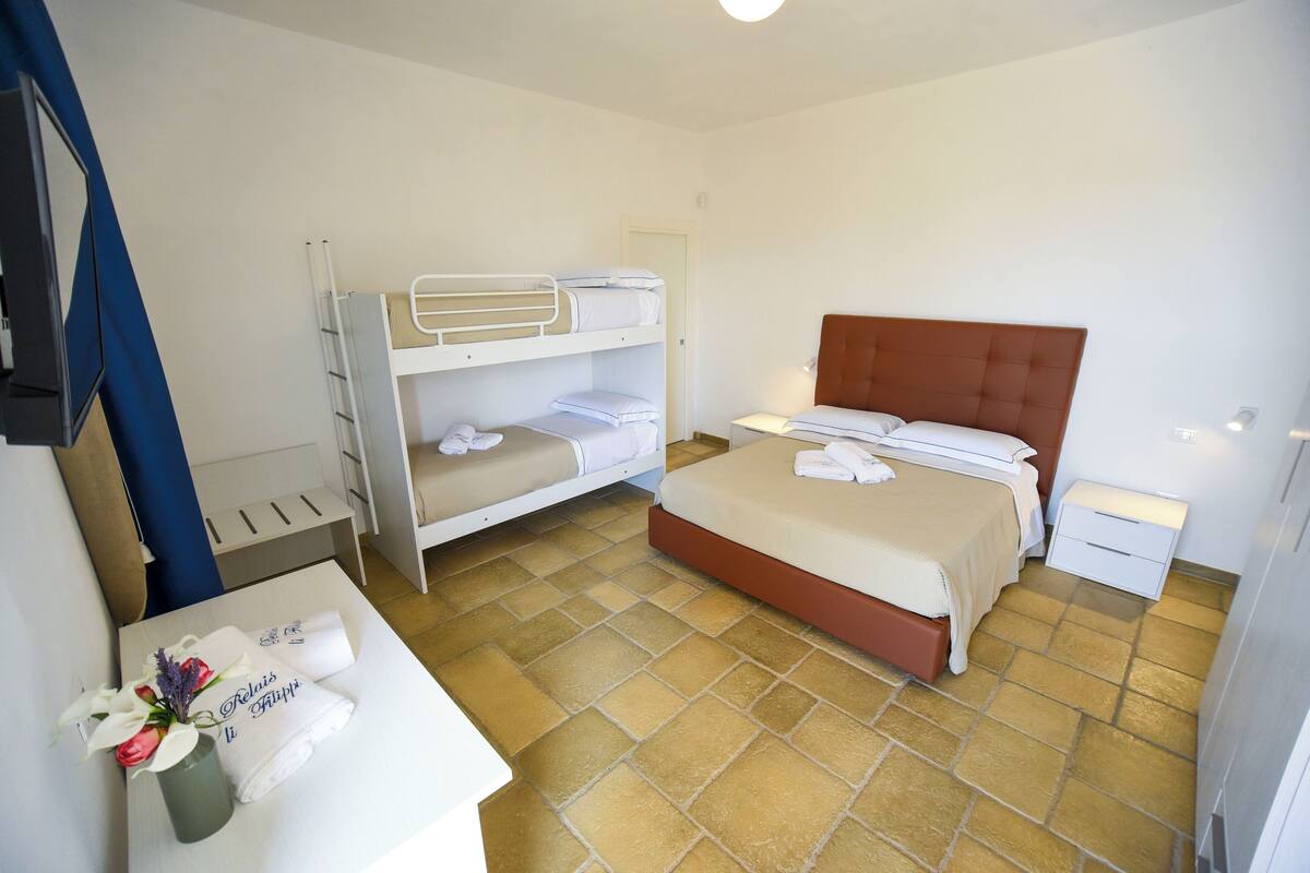 A hotel room with a bunk bed, a single bed, a bedside table with a lamp, and a small desk with a vase of flowers.