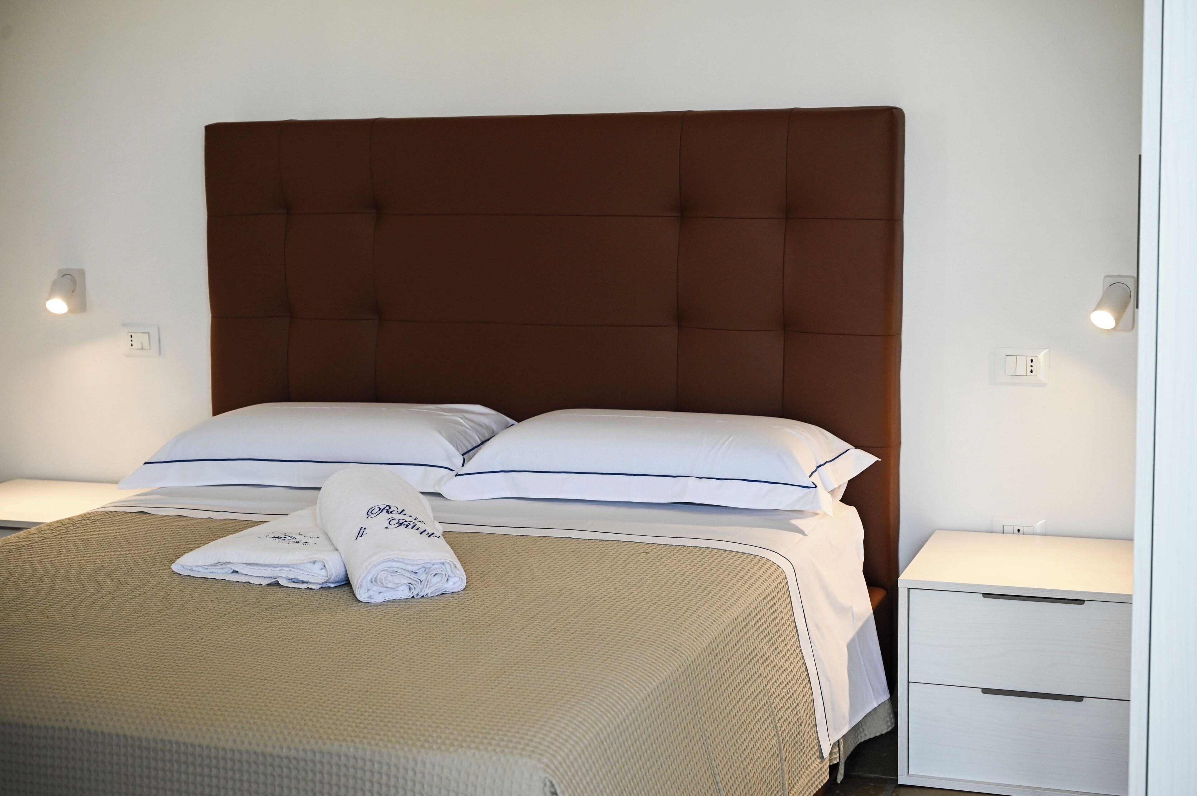 Un lit bien fait, avec une tête de lit marron, des oreillers blancs et une serviette pliée posée sur le lit. Il y a deux tables de chevet, l’une avec une lampe et l’autre avec un tiroir.
