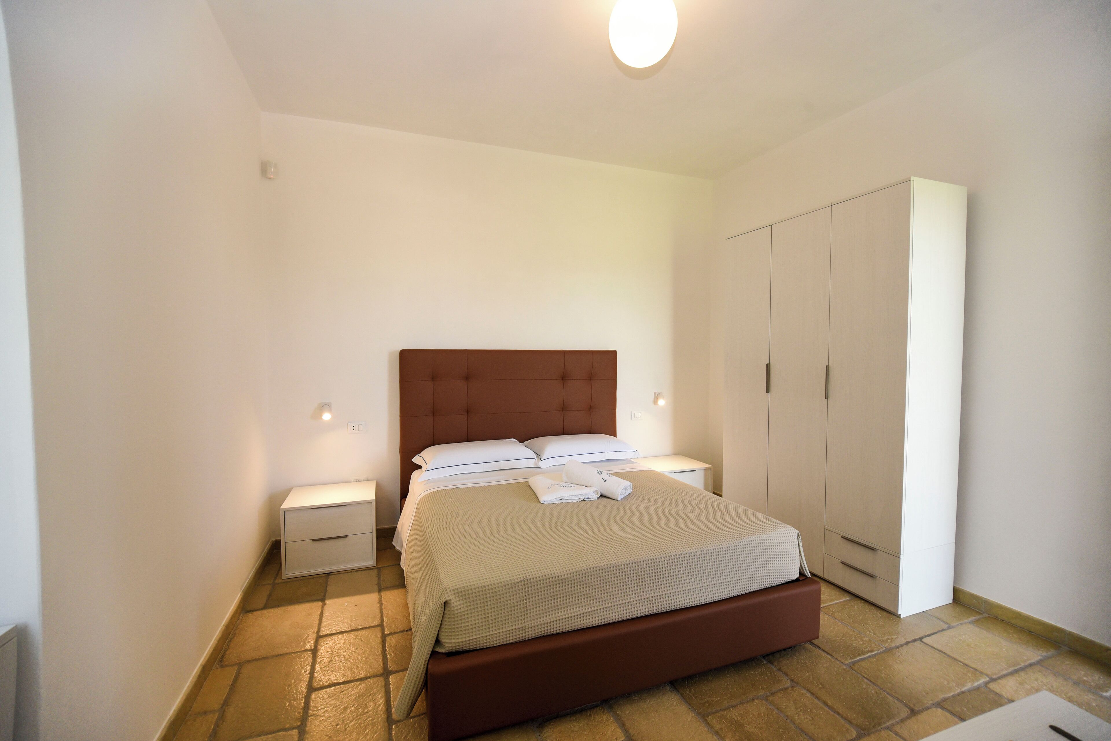 A bedroom with a bed, nightstands, a wardrobe, and a tiled floor.