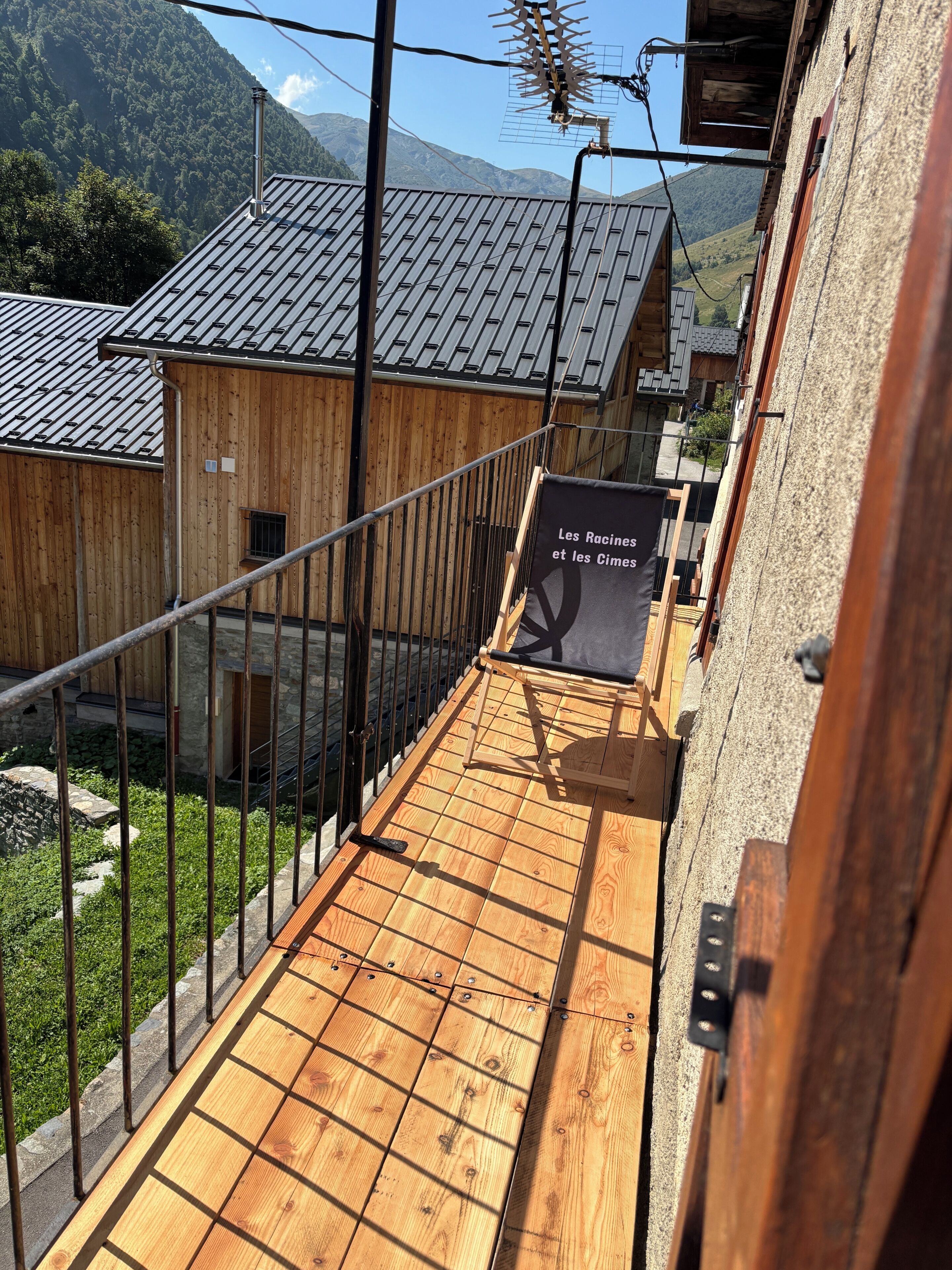 A wooden deck with a metal railing, a sign reading 'Les Racines et les Cimes,' and a mountainous backdrop.