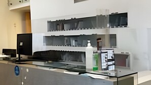 Ein Empfangsbereich mit Glastrennwand, einem Computer und einer kleinen Pflanze. Darüber befinden sich Regale mit verschiedenen Gegenständen.