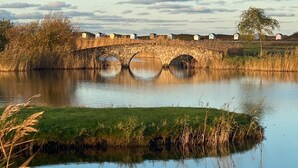Un pont de pierre enjambe une rivière paisible, bordée de rives verdoyantes, avec de petites maisons en arrière-plan.