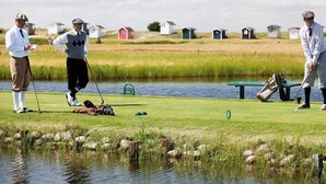 Un parcours de golf avec un étang, des chaises vertes et un drapeau sur un mât.