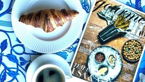 Une table avec un croissant, une tasse de café, une assiette ornée d’un arrangement floral et un magazine.