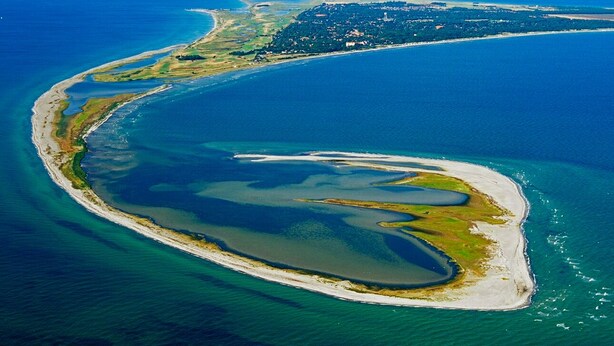Vue aérienne d’une zone côtière avec une baie en forme de courbe, des eaux d’un bleu limpide et une petite île couverte de végétation.
