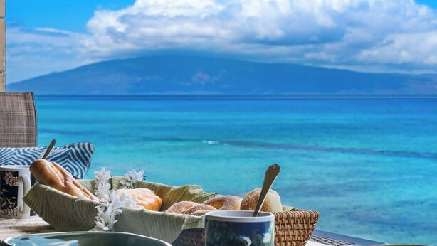 A breakfast setup with bread, pastries, and a mug on a table with a view of the ocean and a mountain in the background. - Top Notch Rental Experience Awaits! (Lahaina)