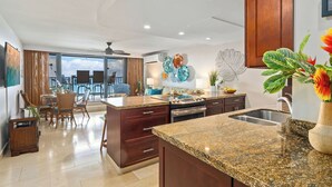 A modern kitchen with granite countertops, wooden cabinets, and a large island. There is a dining area with wicker chairs and a view of the ocean. - Top Notch Rental Experience Awaits! (Lahaina)