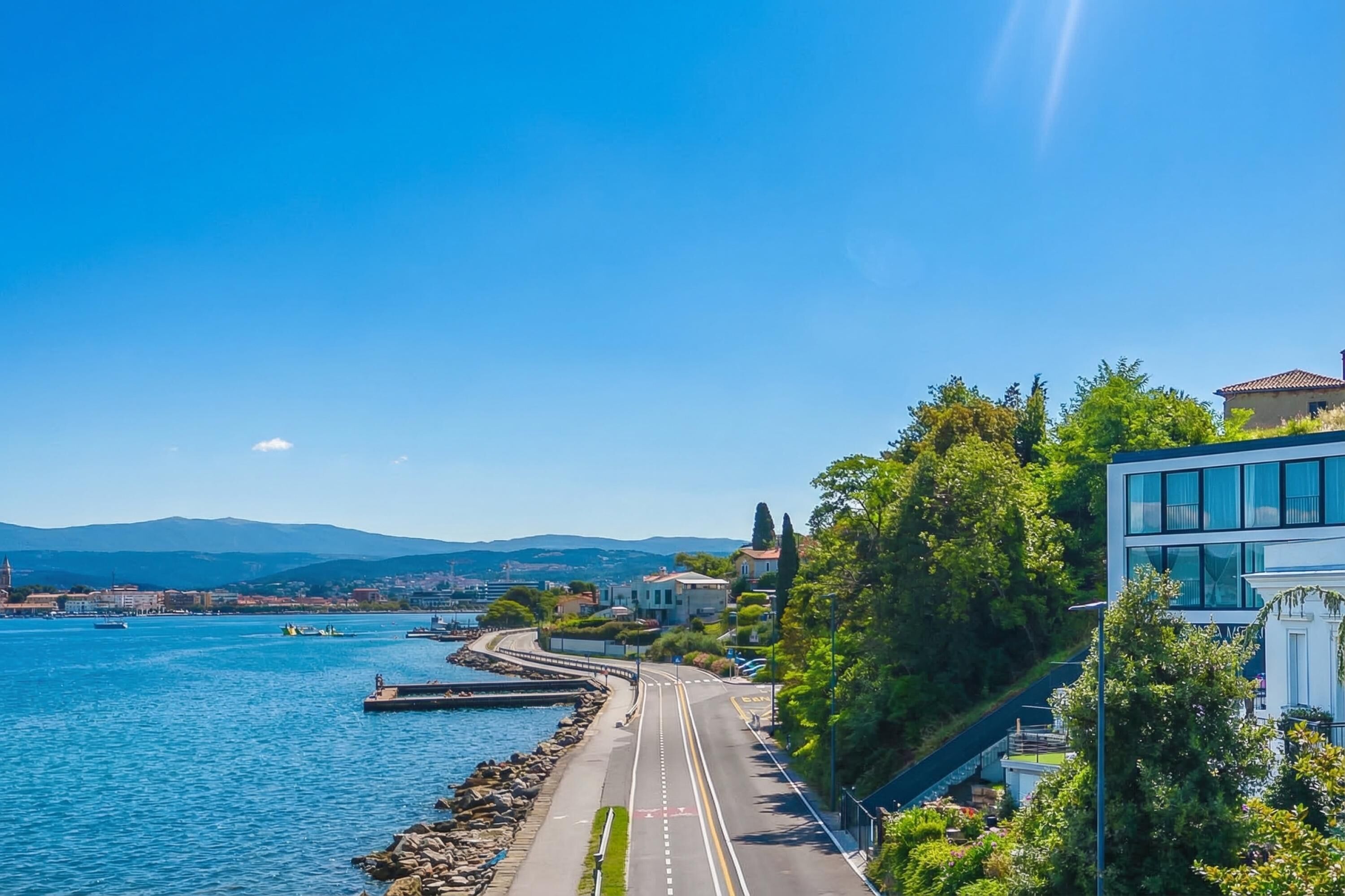 A coastal road with a clear view of the sea, a modern building, and a distant cityscape.