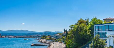 A coastal road with a clear view of the sea, a modern building, and a distant cityscape.