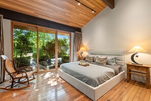 A modern bedroom with a large bed, wooden flooring, and a view of greenery through sliding glass doors. - Waterfront Grandeur in Kelowna (Kelowna)