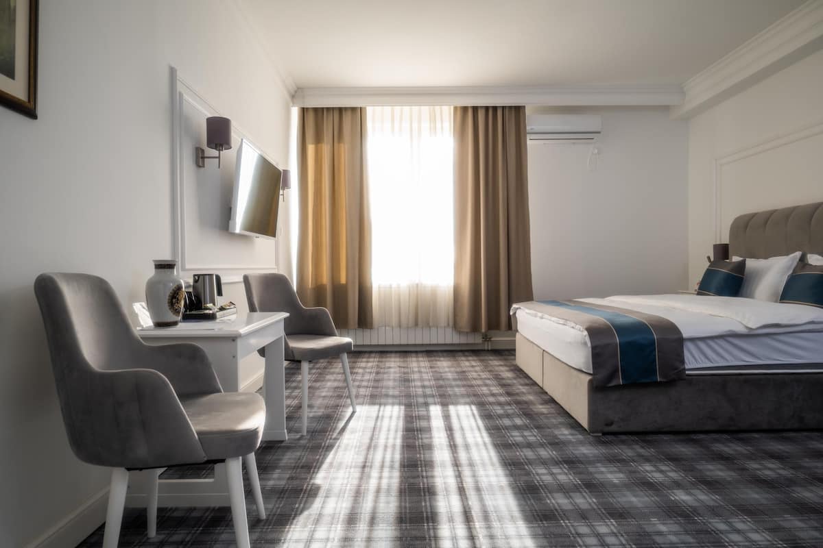 Une chambre d’hôtel comprenant un lit, un bureau, deux chaises, une télévision et une fenêtre avec des rideaux.