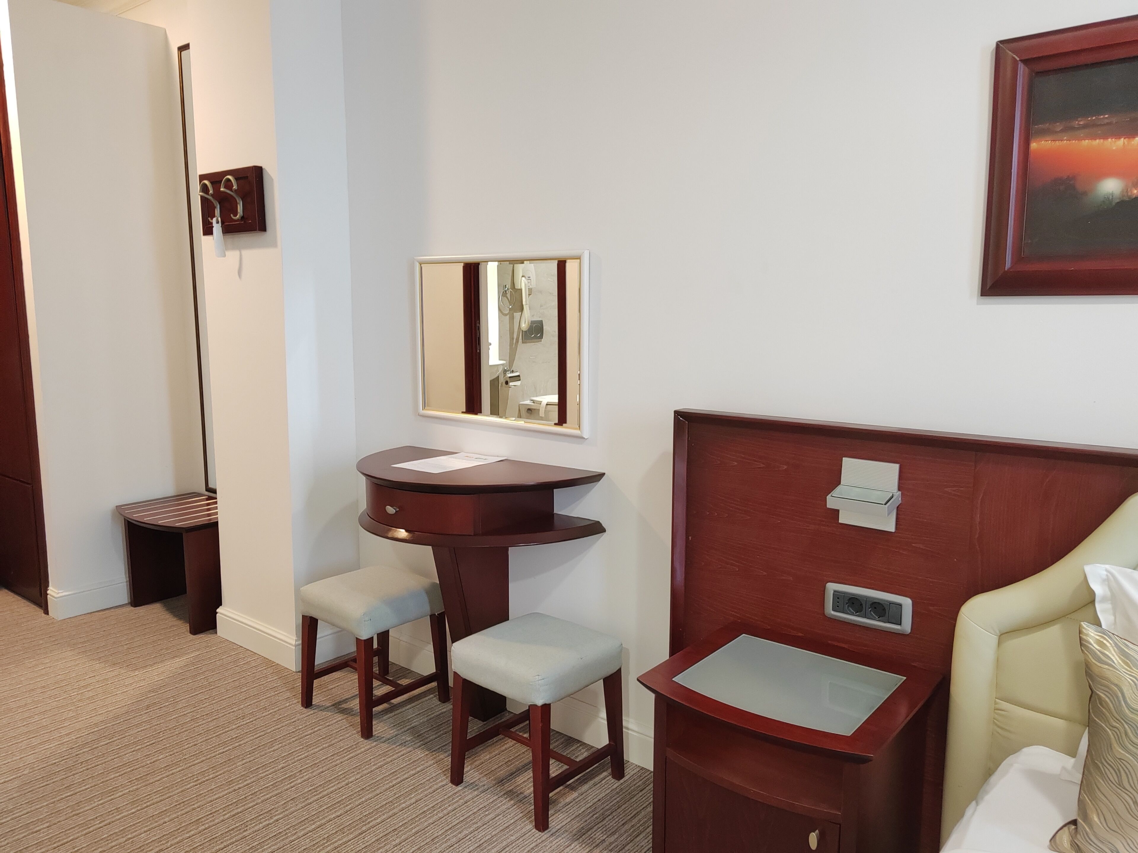 Habitación de hotel con cama, mesita de noche con lámpara, un pequeño escritorio con silla y un espejo en la pared.