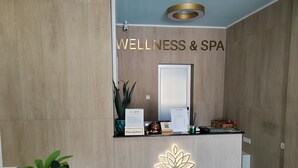A reception desk with a sign reading 'WELLNESS & SPA,' a mirror, a plant, a sign with 'Reception,' and a lotus light fixture. - Mio City Hotel (Kula)