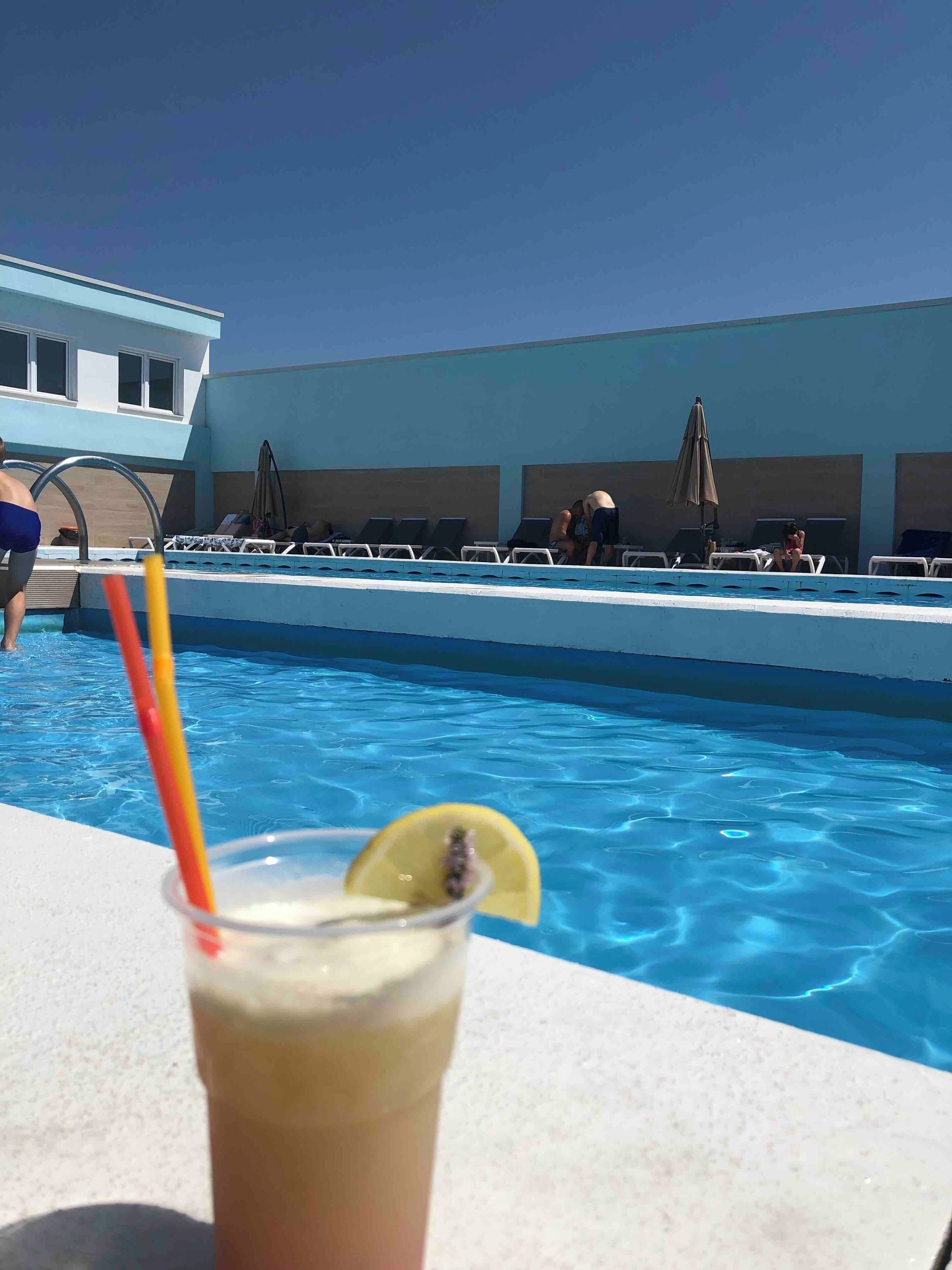 A pool area with lounge chairs and umbrellas, a clear blue pool, and a drink with a lemon slice.
