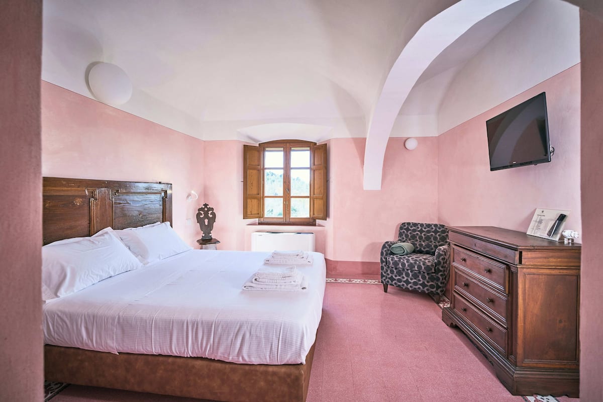 A bedroom with a large bed, a dresser, a chair, a TV, and a window with wooden shutters.