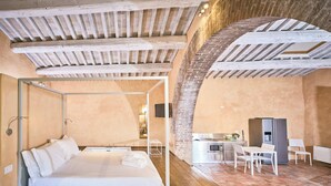 A modern bedroom with a bed, a small table, and a chair. There is a kitchen area with a refrigerator and a sink. The room has exposed wooden beams on the ceiling and brick walls. - Sant'Anna in Camprena Agriturismo (Pienza)