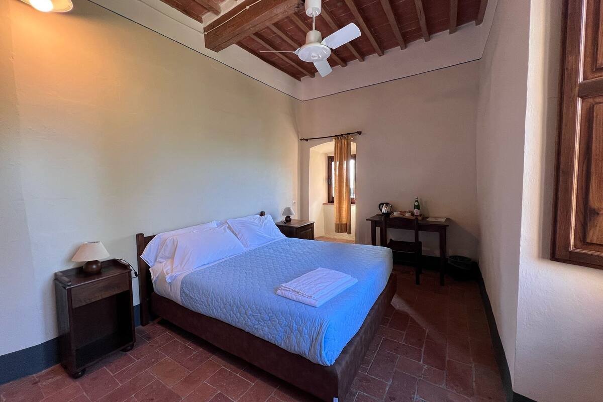 A bedroom with a bed, a nightstand, a desk, a ceiling fan, and a window with curtains.