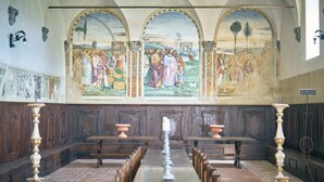 A church interior with a long wooden pews, a central aisle, and a fresco mural on the wall depicting a religious scene. - Sant'Anna in Camprena Agriturismo (Pienza)