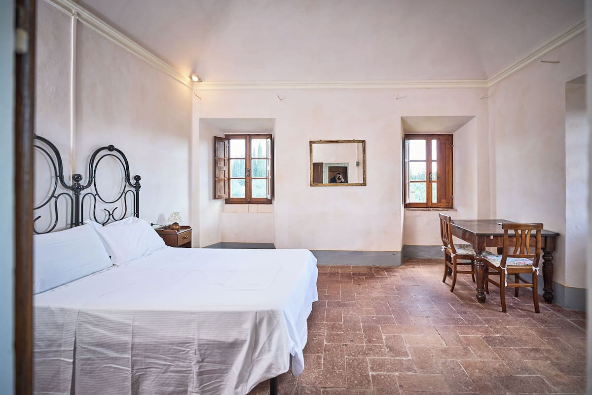 A bedroom with a bed, a wooden table with chairs, and two windows.