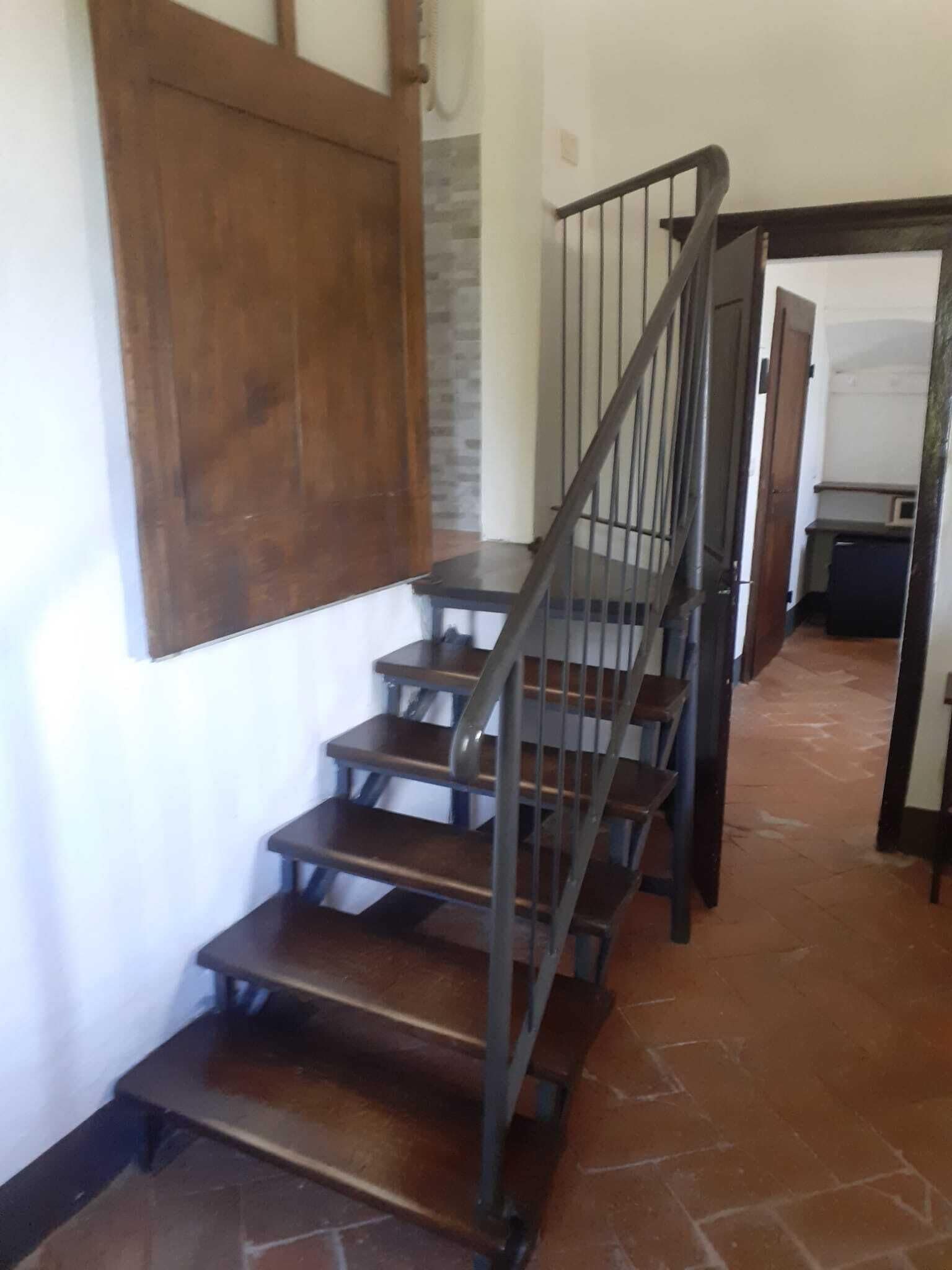 Una scala con corrimano in legno e balaustre metalliche, che conduce a una stanza con pavimento piastrellato e una porta sullo sfondo.
