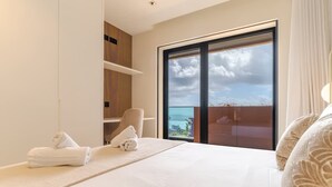 Habitación de hotel moderna con una cama grande, una silla, una mesita y vistas al mar a través de un ventanal.