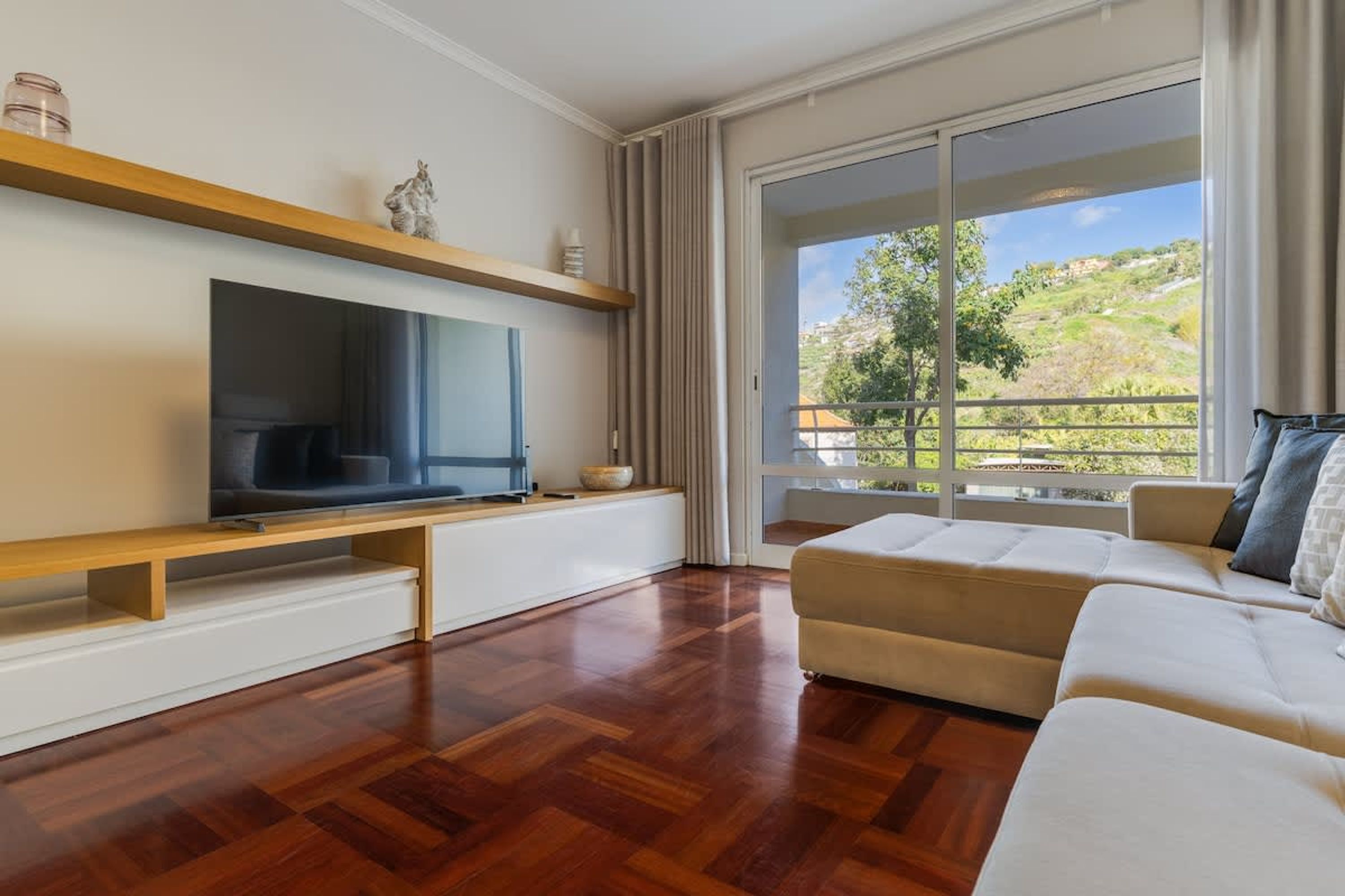 A modern living room with a flat-screen TV, wooden shelves, a beige sofa, and a large sliding glass door leading to a balcony with a view of greenery.
