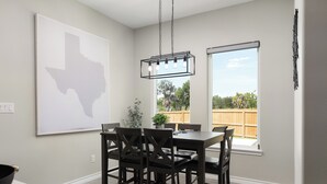 A dining area with a table and chairs, a hanging light fixture, a window with a view of the outdoors, and a wall-mounted artwork.