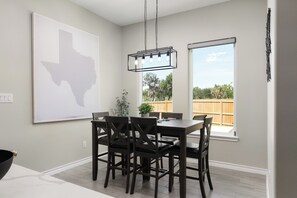 A dining area with a table and chairs, a hanging light fixture, a window with a view of the outdoors, and a wall-mounted artwork.
